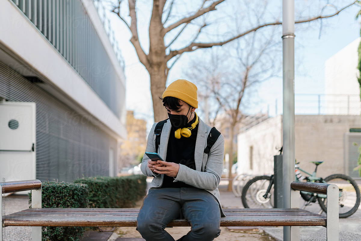 Serious man in mask using smartphone on bench on street
