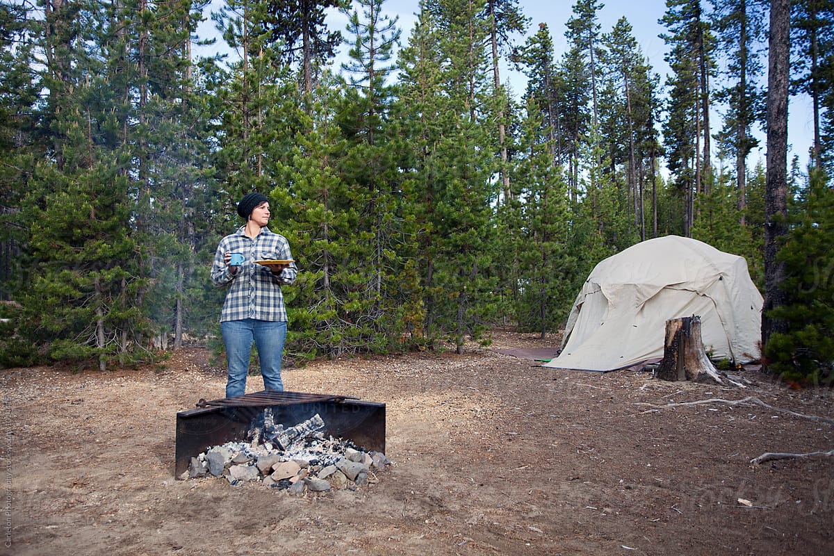 Woman in plaid flannel shirt and jeans has breakfast next to a campfire