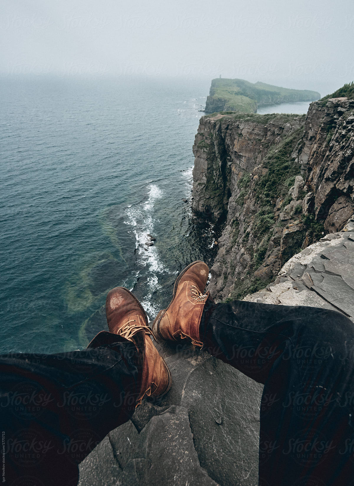 Man in leather vintage boots sitting on the edge of a cliff and looking at the sea.