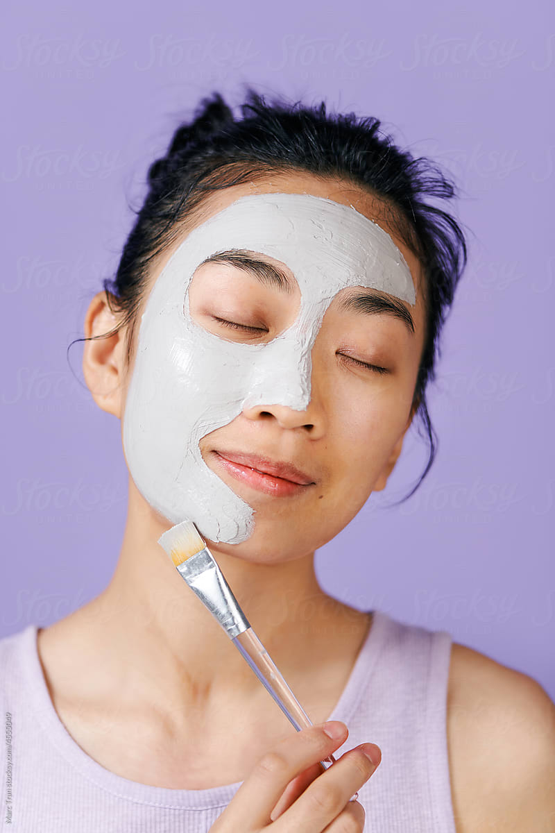 Asian woman applies clay mask on face with cosmetic brush