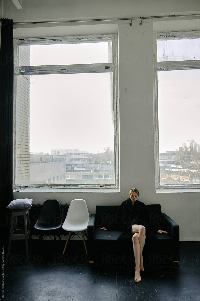 artistic photo of a girl in a black long coat sitting against the background of large windows