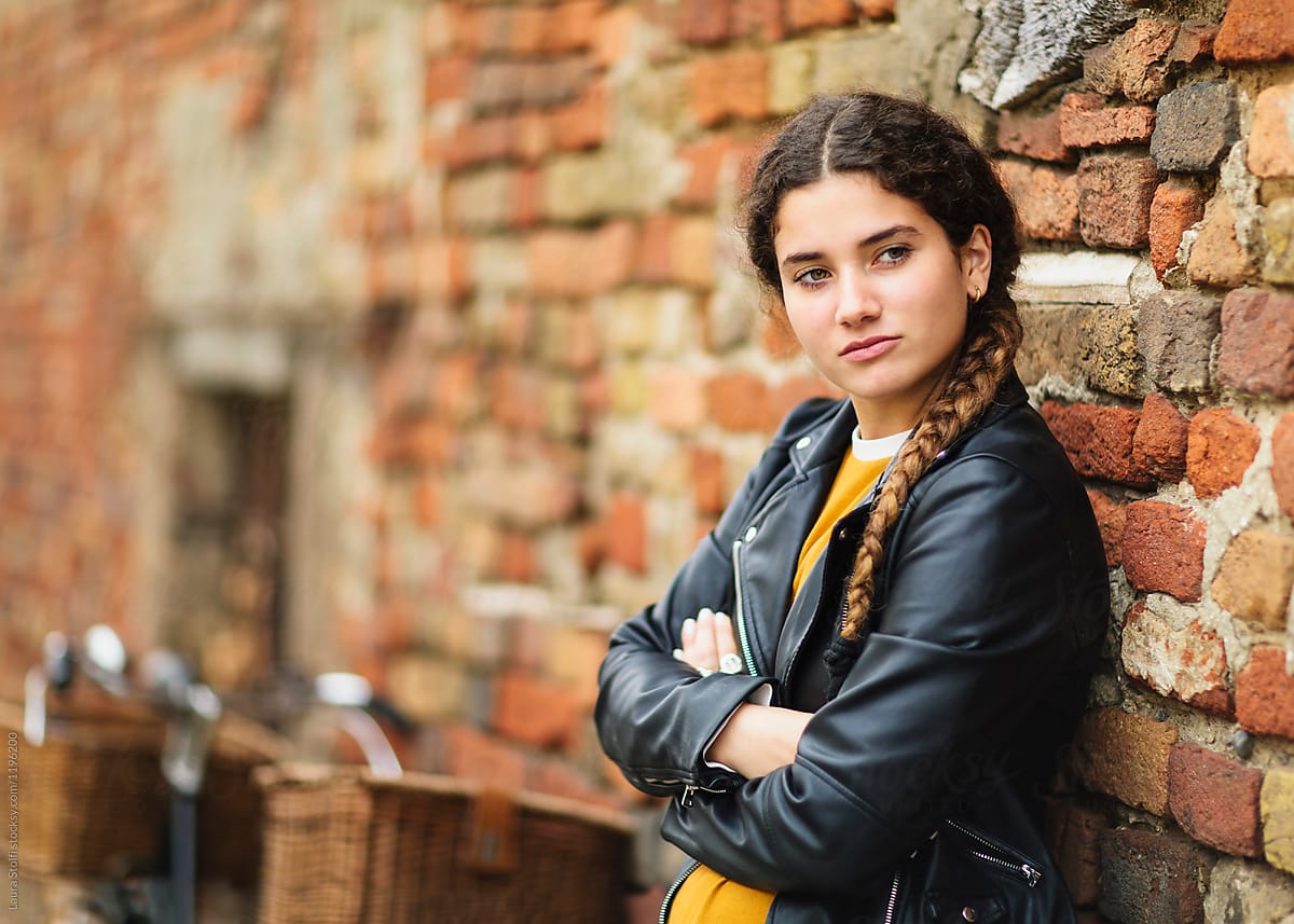 Young girl wearing motorbike jacket leans her back against ancient brick wall in italian street