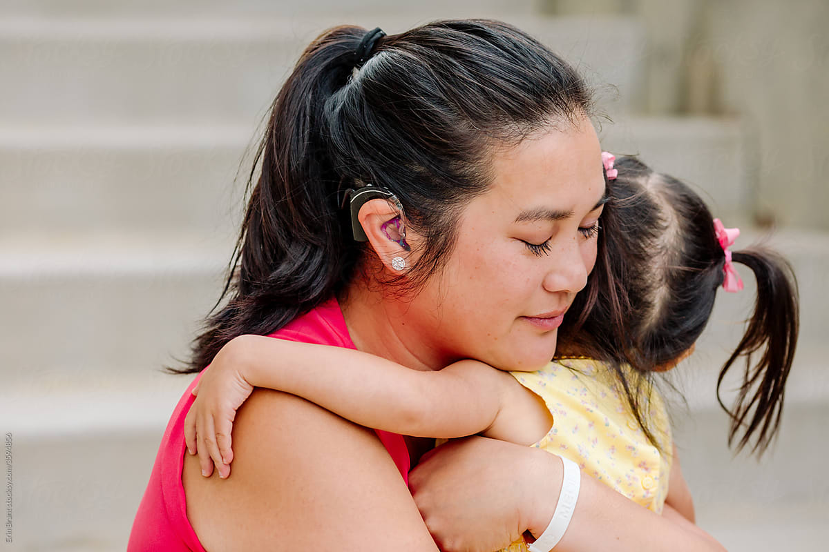 Beautiful Asian mom with cochlear implant hugging young daughter