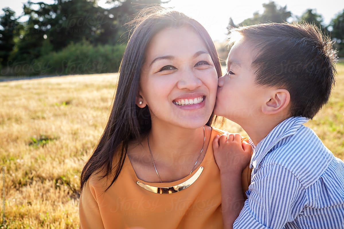 Asian kid kissing his mom on the cheek outdoor in a park