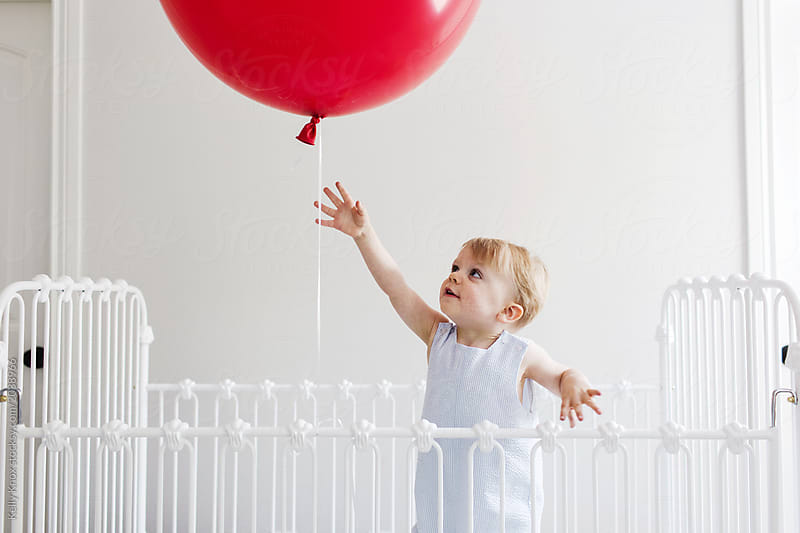 toddler in a crib reaching for a big red birthday balloon