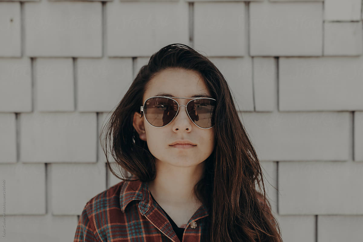 Portrait of young adult woman with long brown hair standing against a white textured wall staring straight at the camera with aviator sunglasses with a serious face