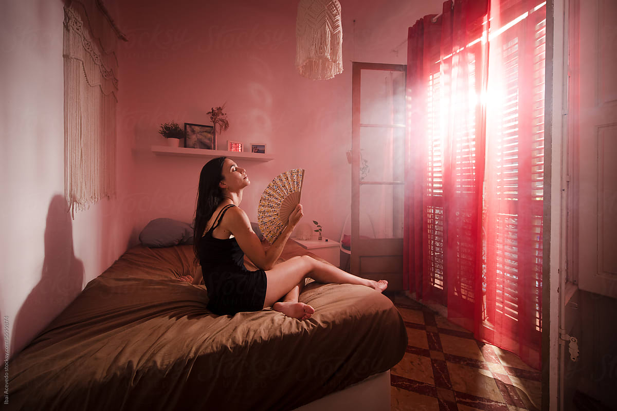 Woman using hand fan sitting on bed at morning