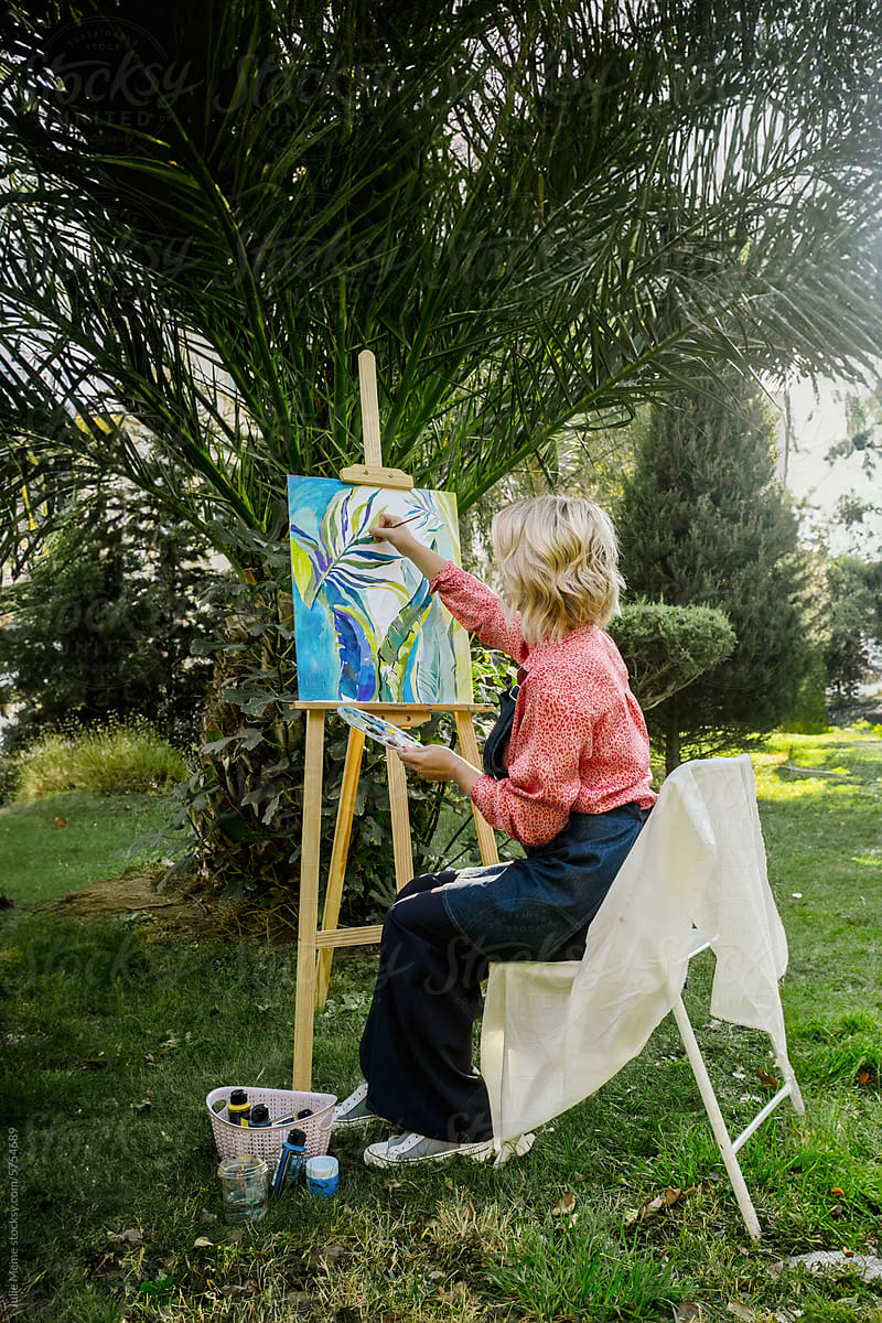 An artist in pink blouse sits with her back turned, paints in nature