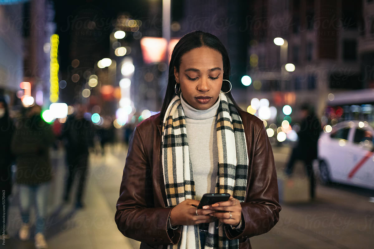 Stylish black woman texting with smartphone in the city at night