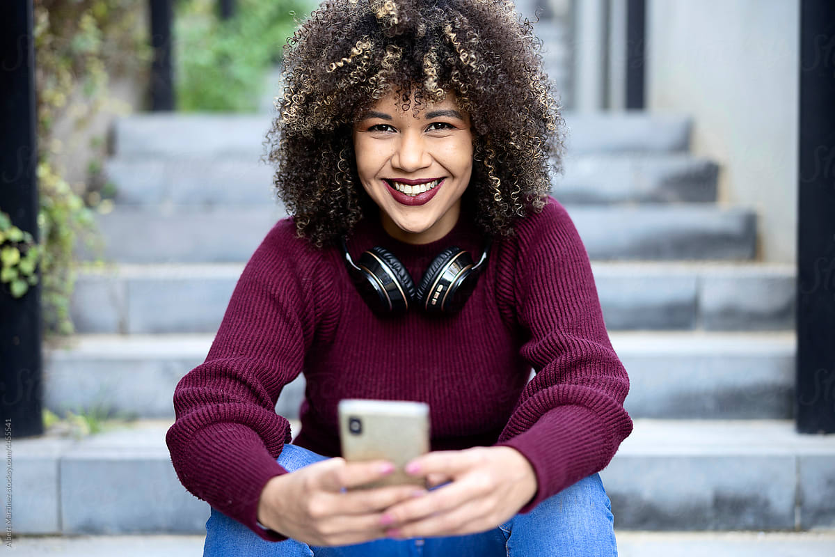 Positive curly haired woman with smartphone in city