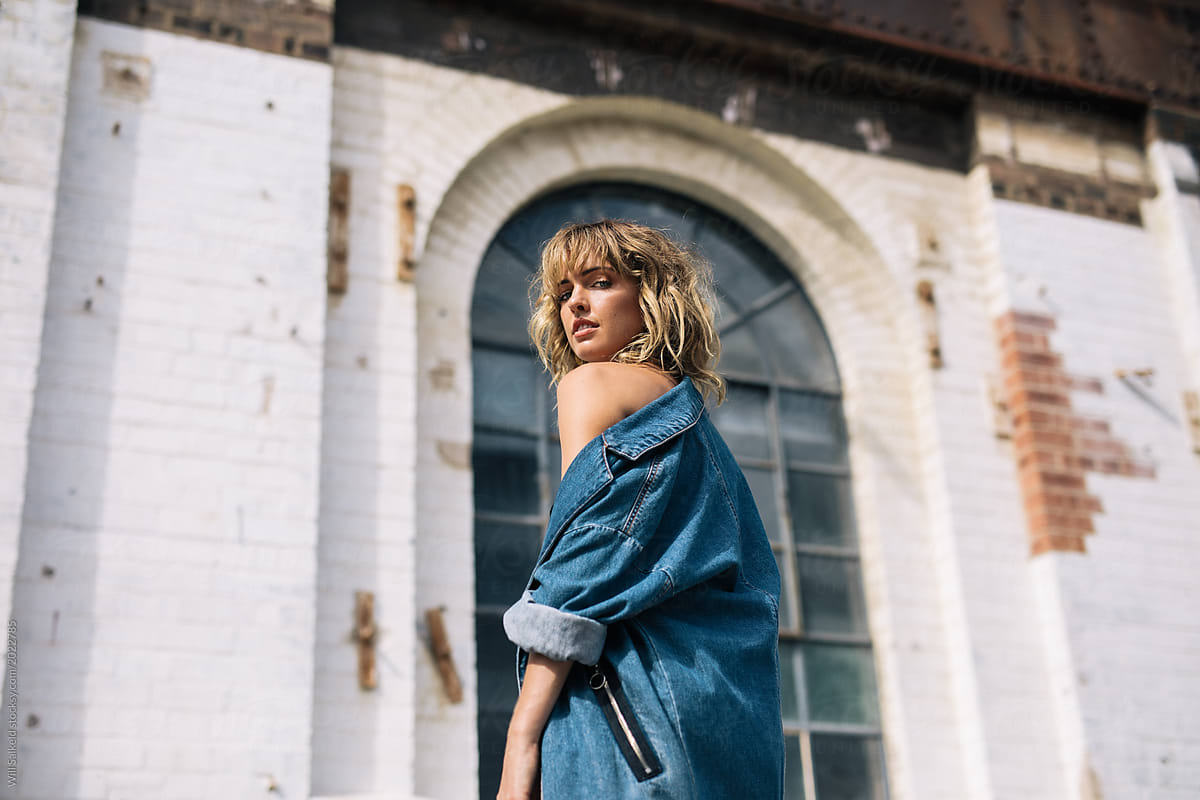 A pretty young blonde girl poses in front of rustic factory style building with denim coat
