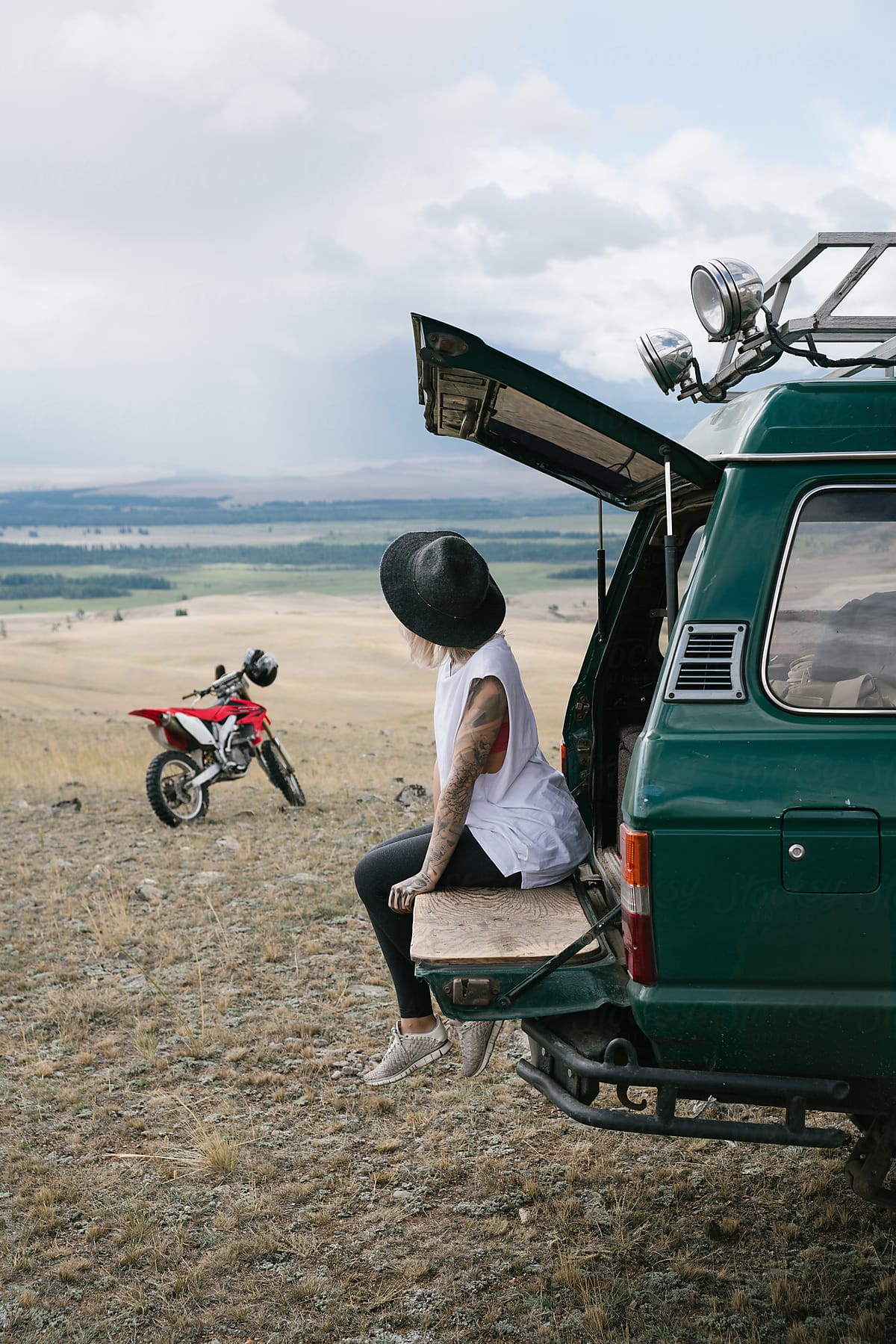 Young tattooed woman sitting in old jeep parked in the wild area surrounded by mountains