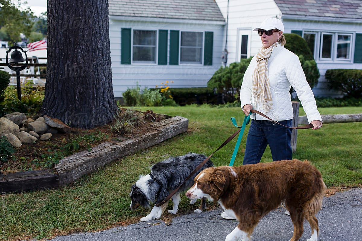 Senior woman holding two dog leashes takes a walk past a house.