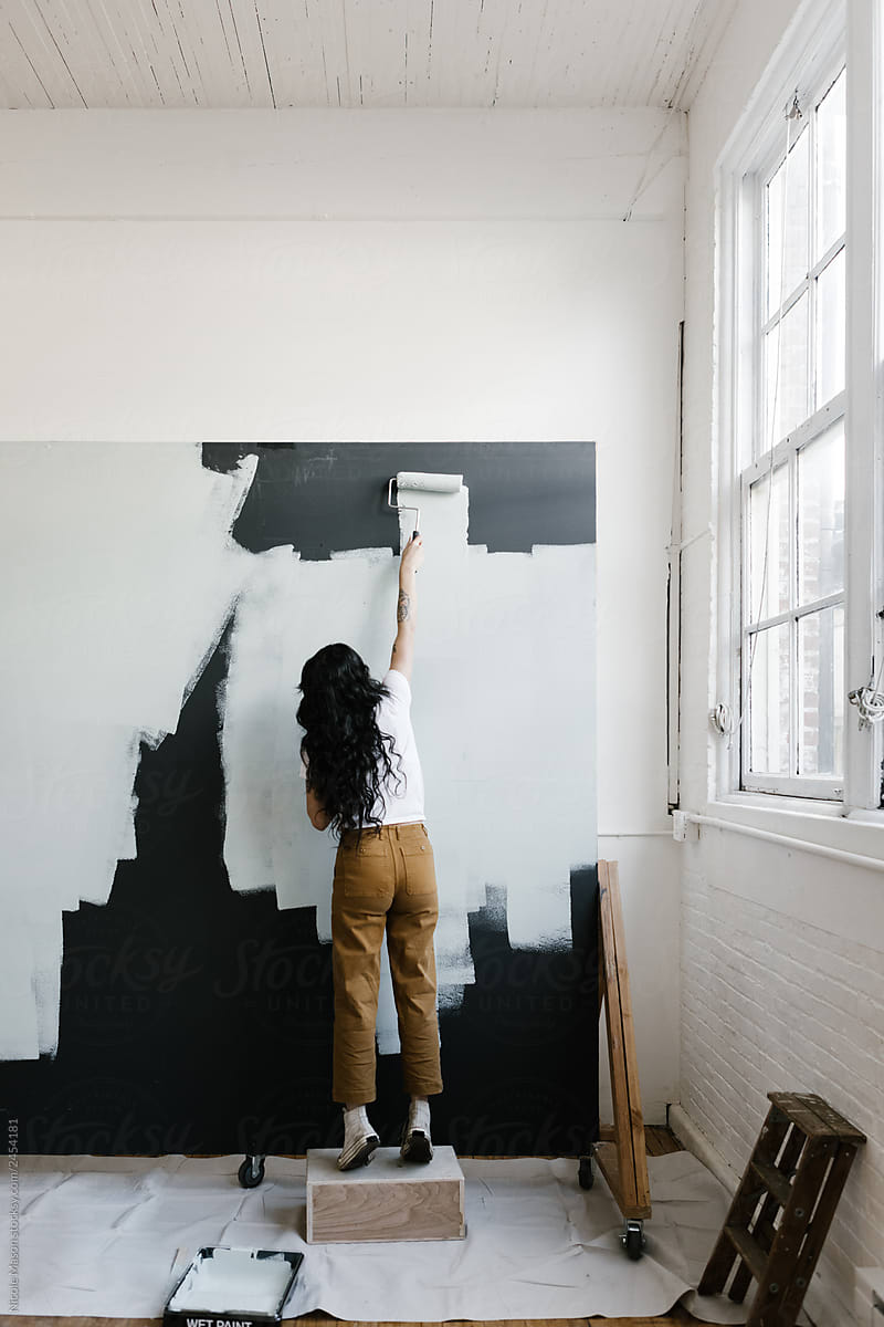 young woman reaching up painting wall in industrial creative studio