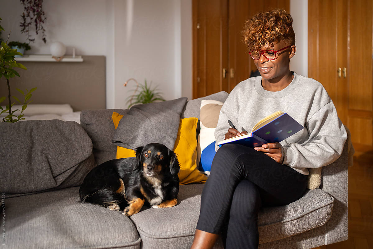 Black Woman With A Book Sitting On The Couch Next To Her Dog.