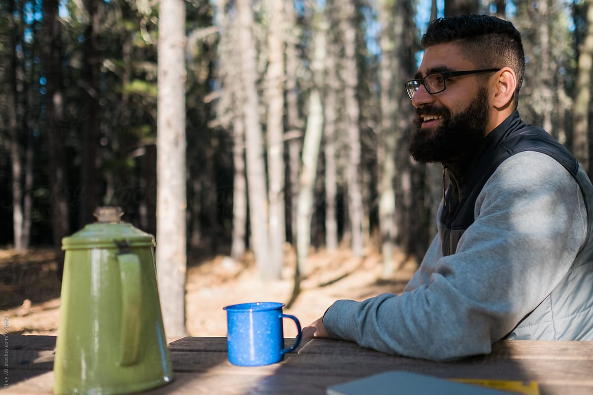 A bearded man smiles while sitting at a picnic table with enameled mugs and a kettle.