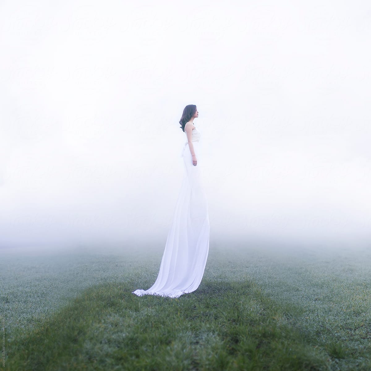 Between sky and earth. Beautiful young woman standing in a field in a long white dress