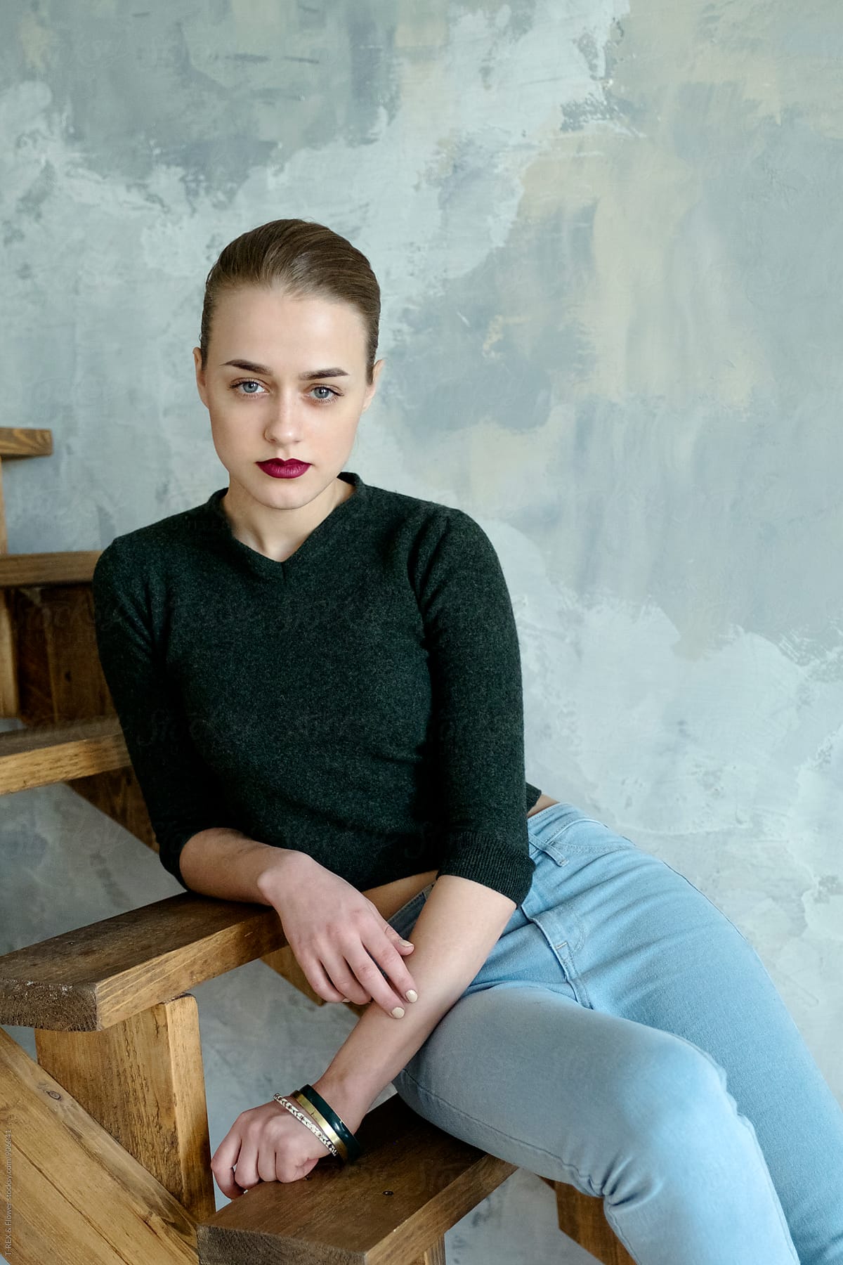 Woman In Jeans And Sweater Sitting On Stairs While Looking At Camera By Stocksy Contributor