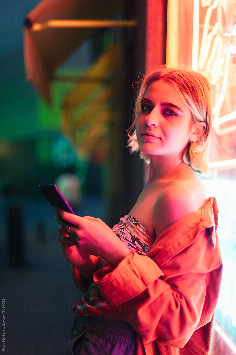 cool woman using smartphone leaning on a neon panel at night