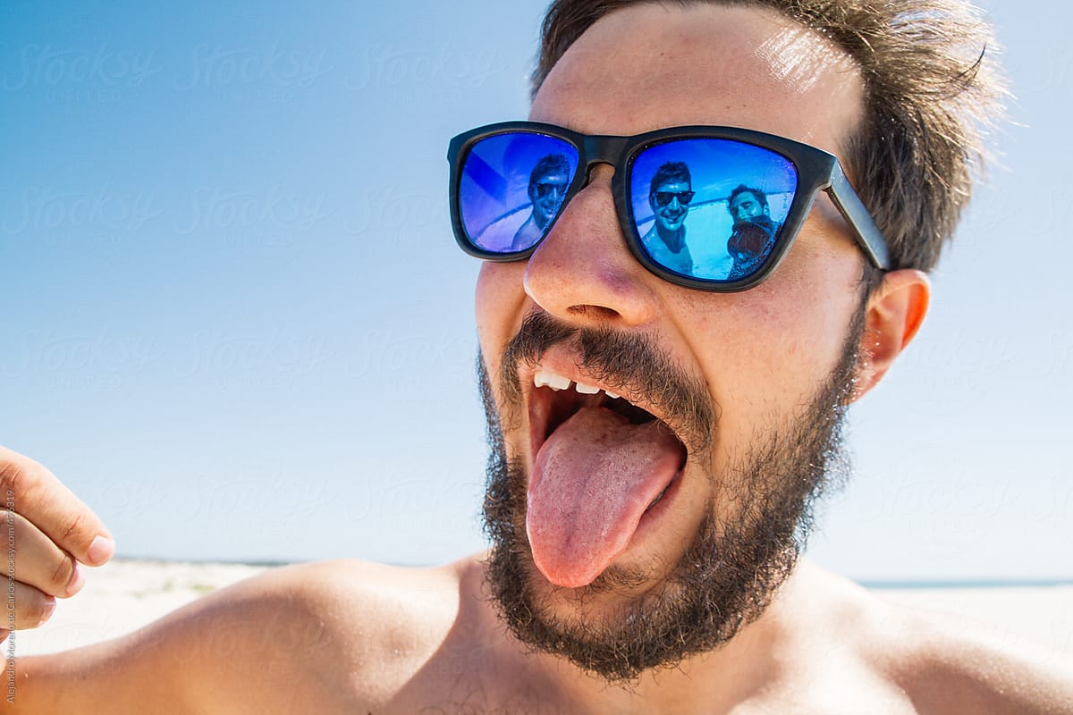 Young Hipster Man Sticking Out Tongue On The Beach With His Friends Showing On The Reflection
