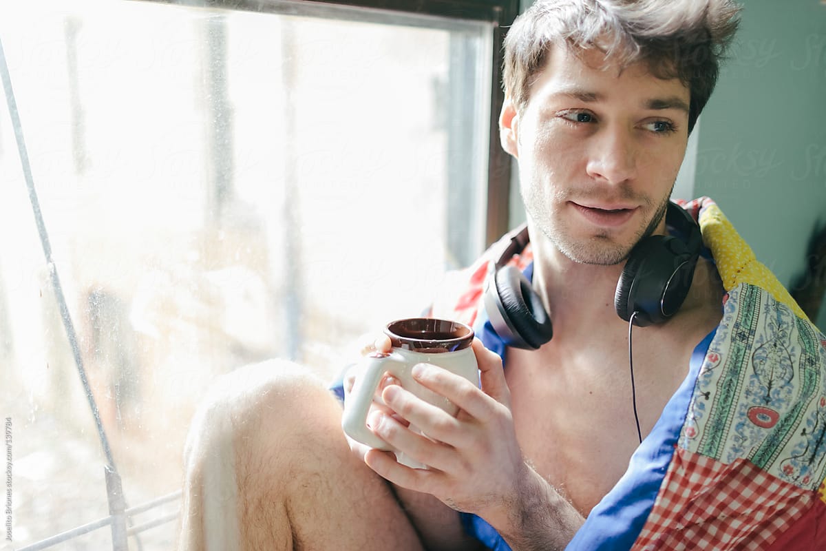 Young Man Waking Up in the Morning Listening to Music and Drinking Coffee by Window with Quilt