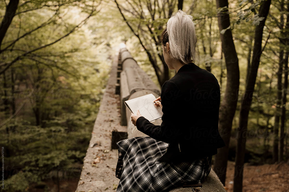 Young woman sitting in old pipeline drawing in notebook in forest during autumn