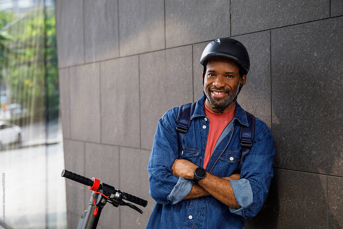 Smiling man wearing helmet leaning on wall by electric push scooter