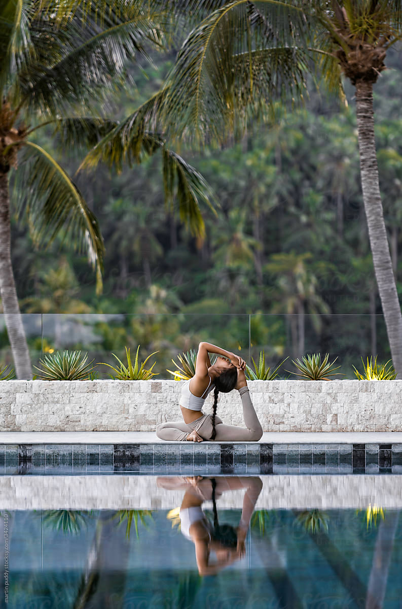 Asian woman practicing yoga on edge of swimming pool in tropical surroundings
