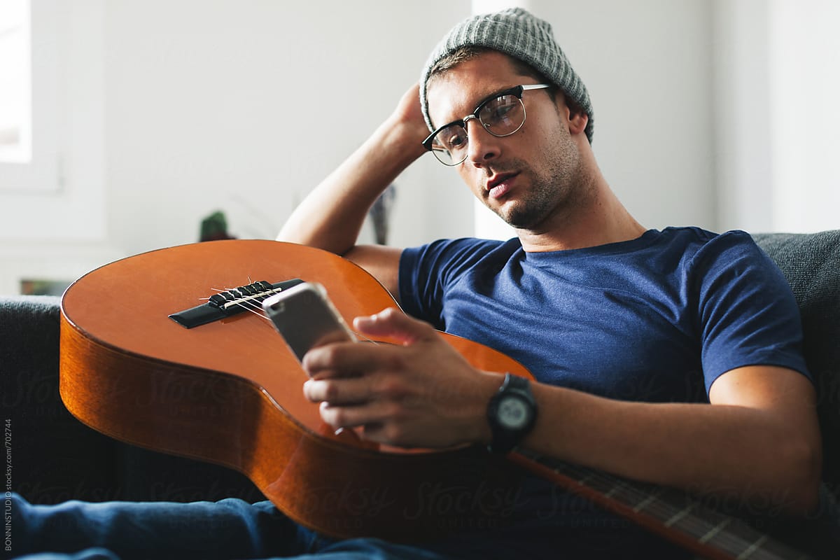 Hipster guitarist man using his phone sitting on the couch at home.