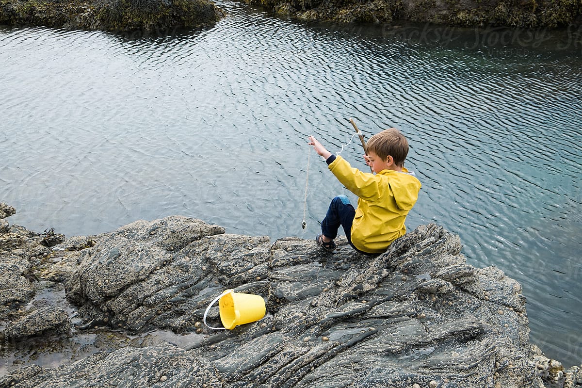 Little Boy Sitting On Rock By The Sea With A Fishing Pole by
