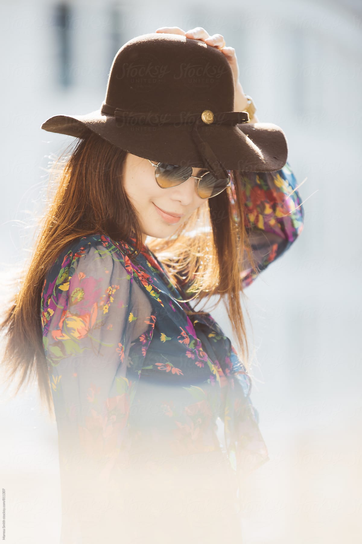 A brunette lady smiling and looking down wearing heart-shaped glasses and a floppy hat