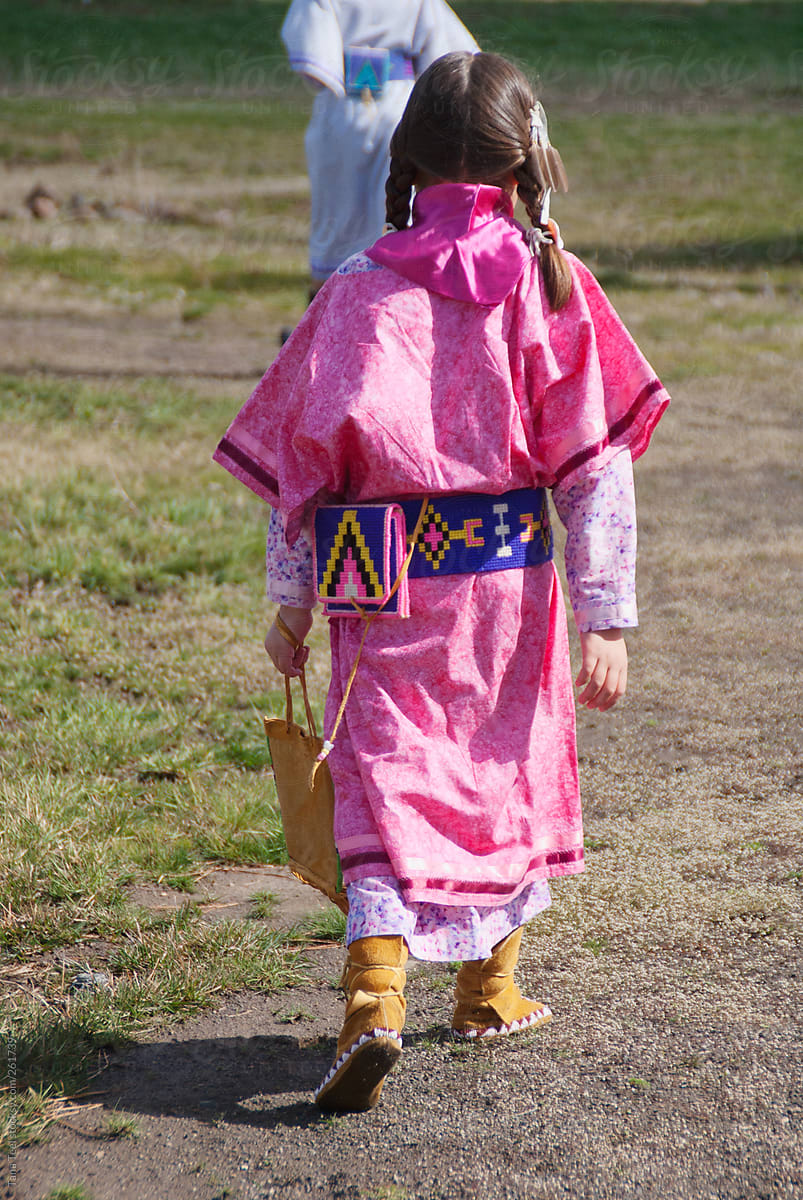 A young Native American girl dressed in traditional clothing walks in a field