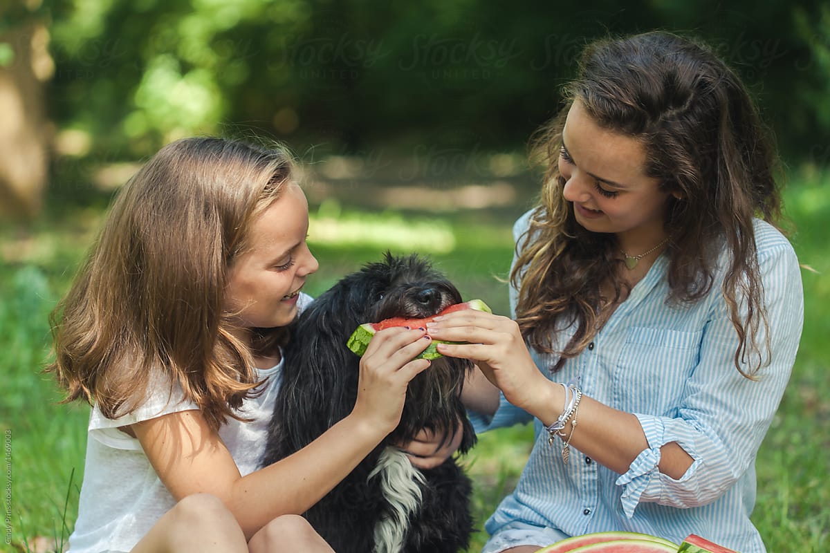 Two smiling girls feeding their dog watermelon