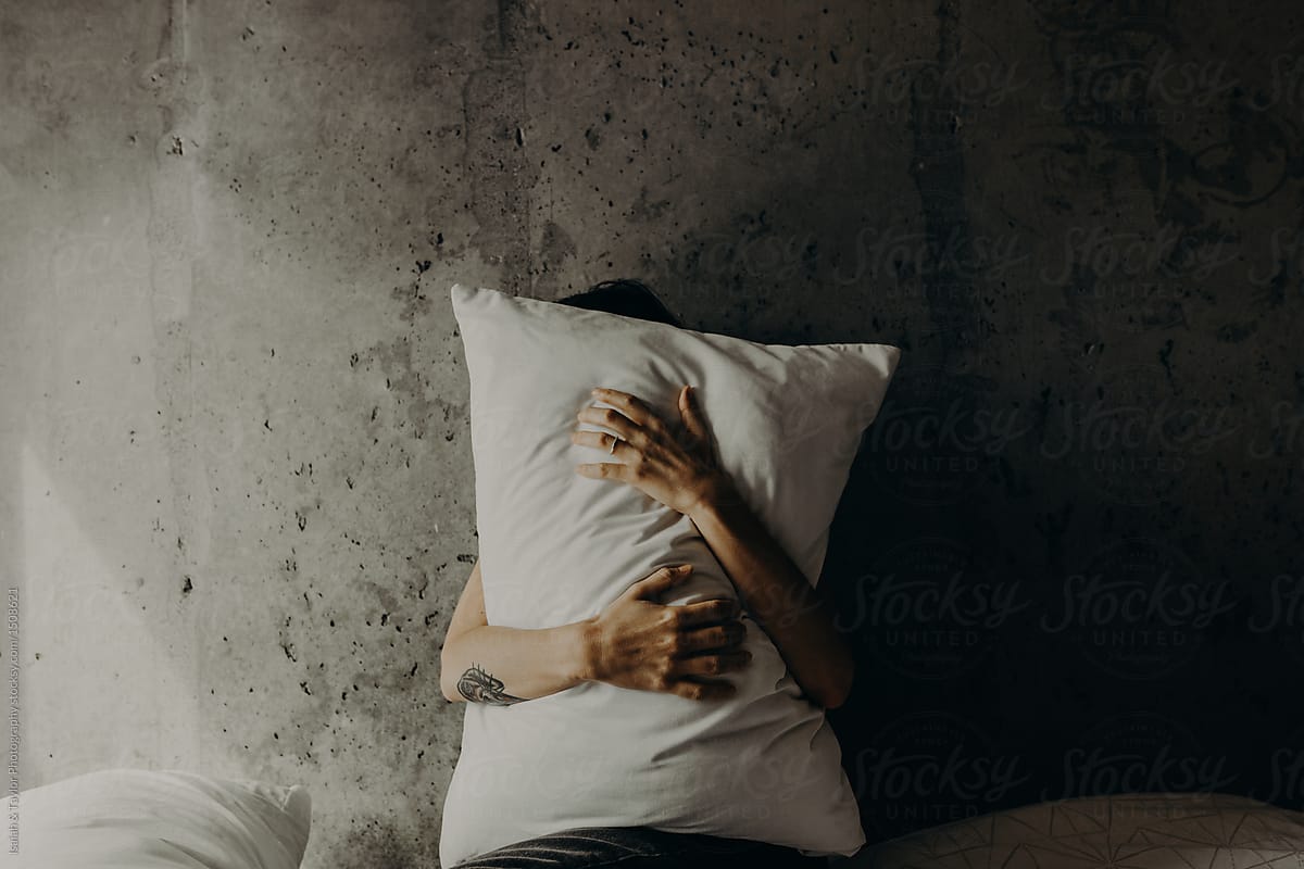 Woman Hiding Behind A Pillow While Sitting Alone On A Bed In A Simple Concrete Loft Bedroom By