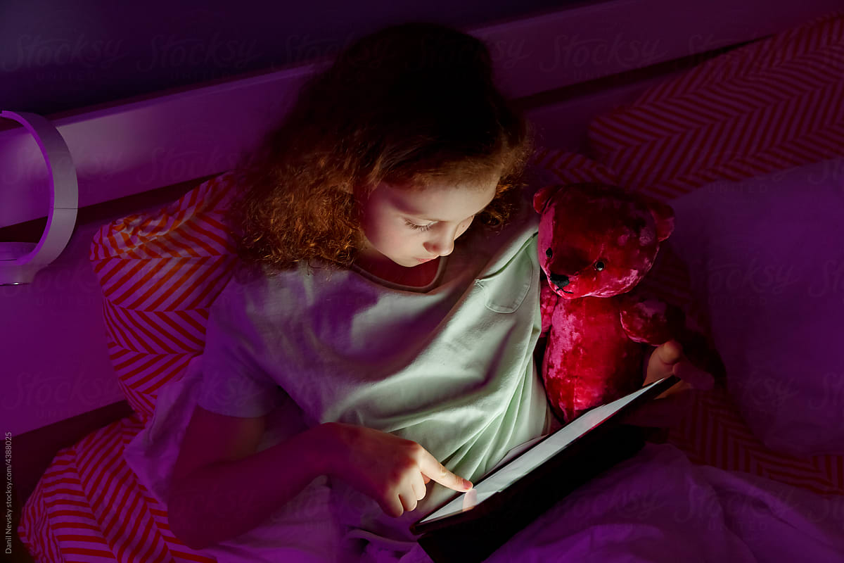 Girl with teddy bear browsing tablet in dark bedroom