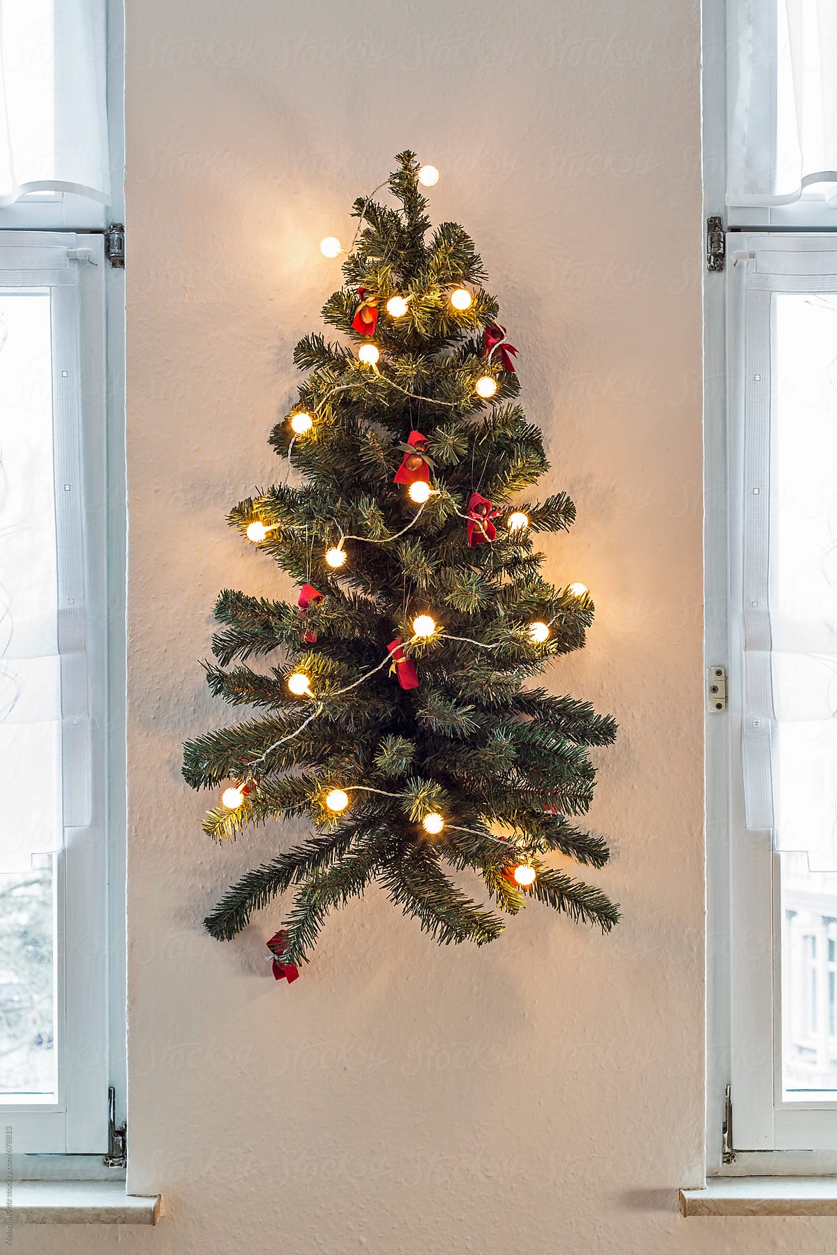 Decorated artificial Christmas tree hangs on a wall between two windows