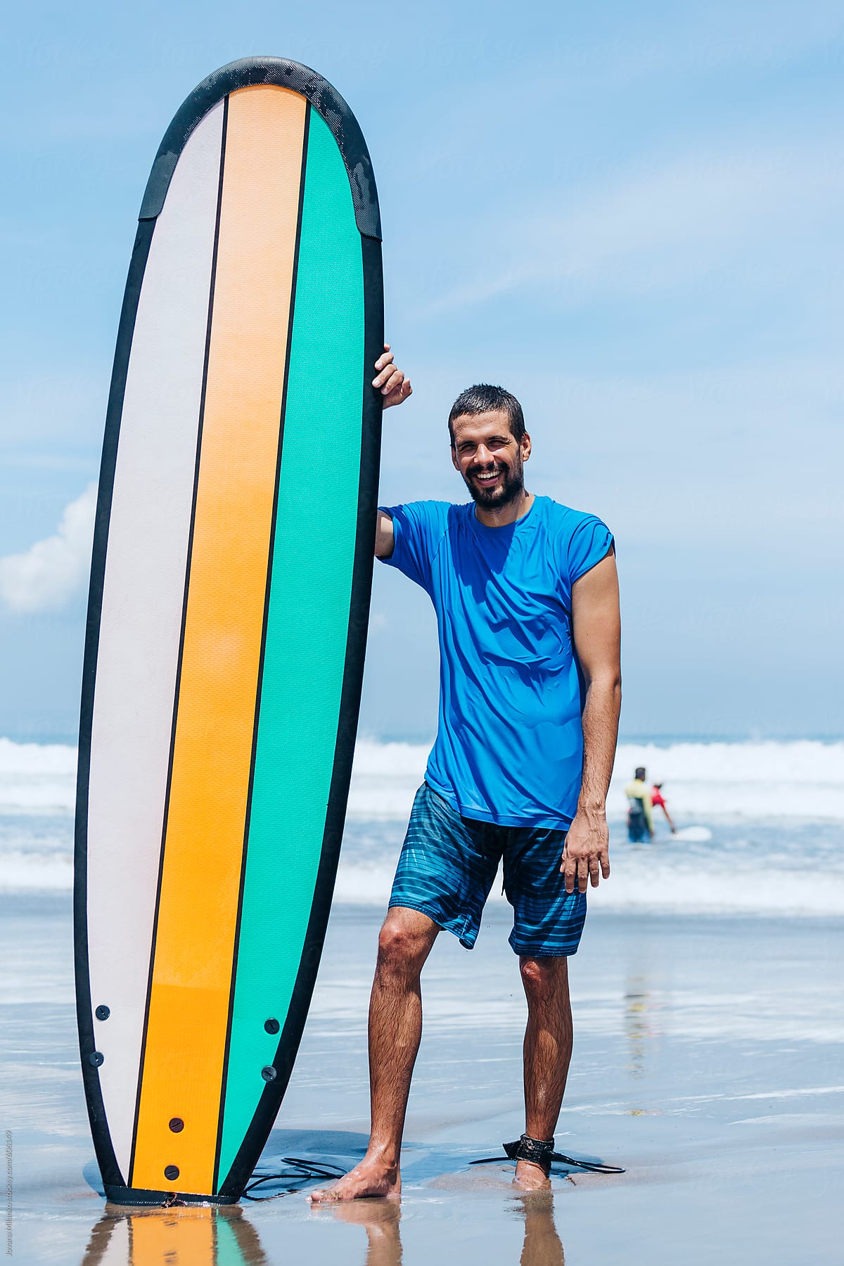 Portrait of a happy surfer guy standing on the beach leaned on a long surfboard