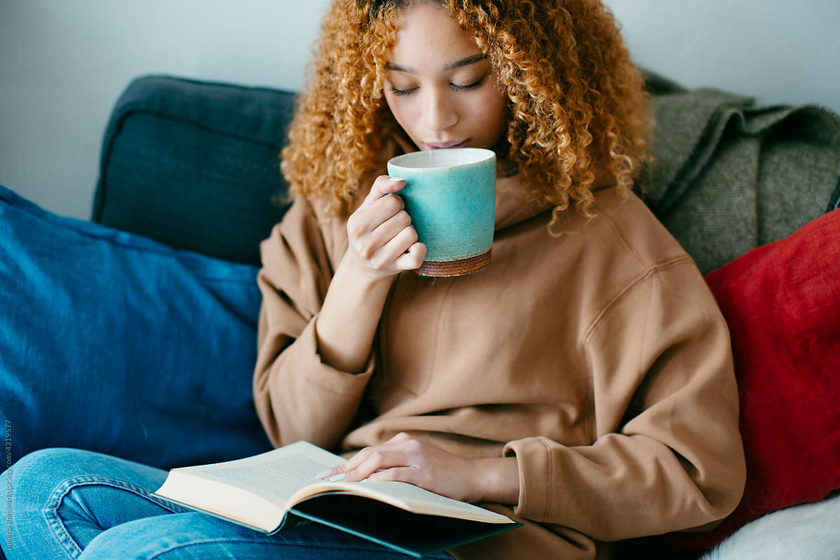 Young woman drinking tea while reading a book on couch