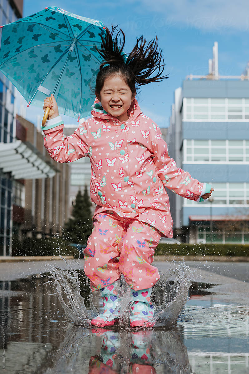 Young girl jumps in a puddle with rain boots and an umbrella.