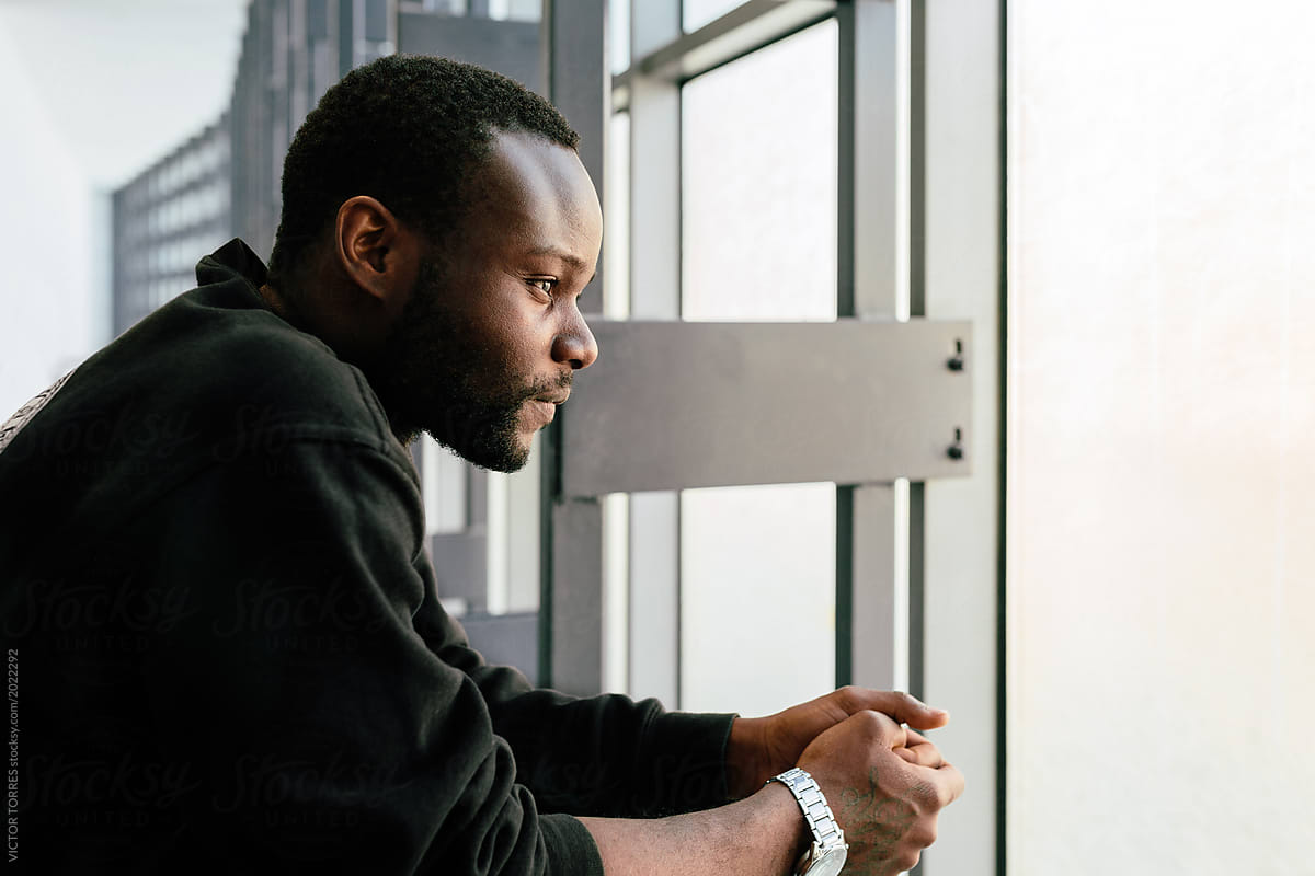 Young black man with pensive expression looking out window by VICTOR