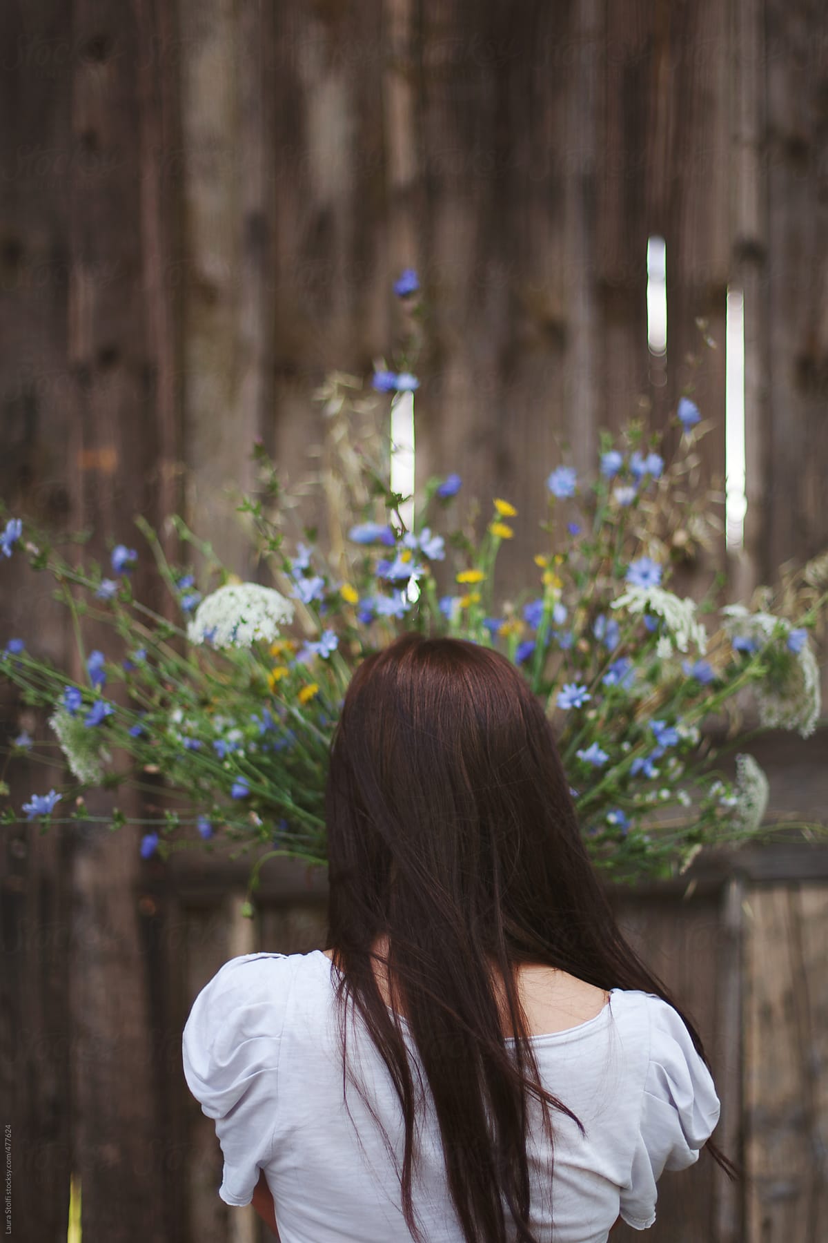 Rear sight of young brunette holding mixed wildflowers bouquet in front of old wooden wall