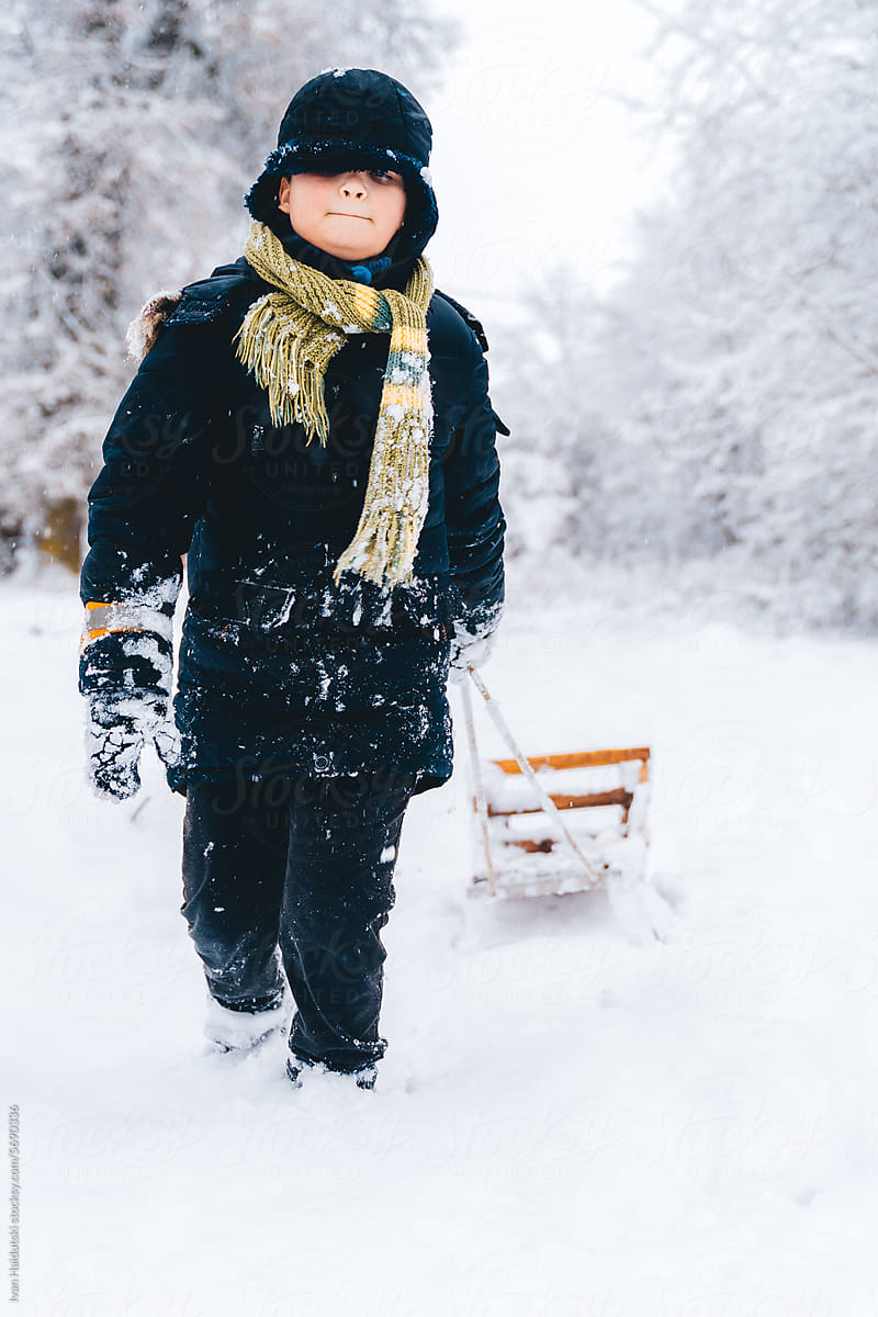 Cute kid boy happily pulls sled along forest road in winter suburbs
