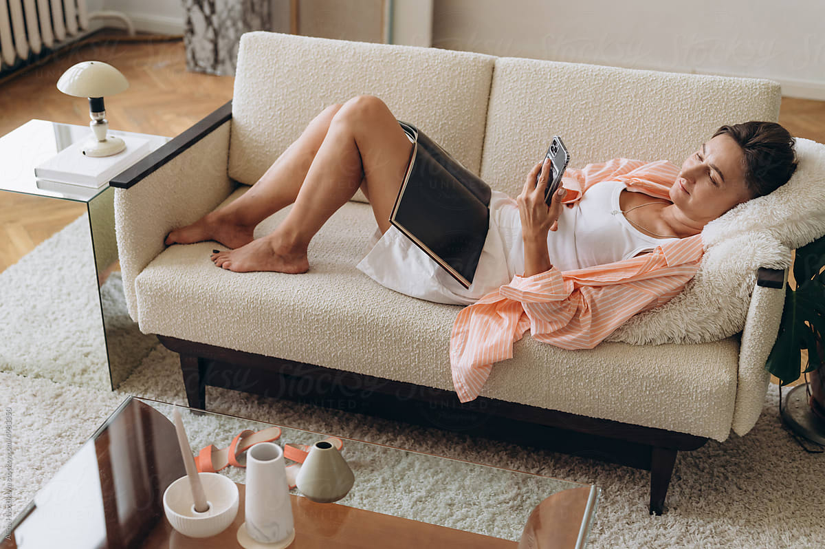 Woman with magazine using smartphone on sofa