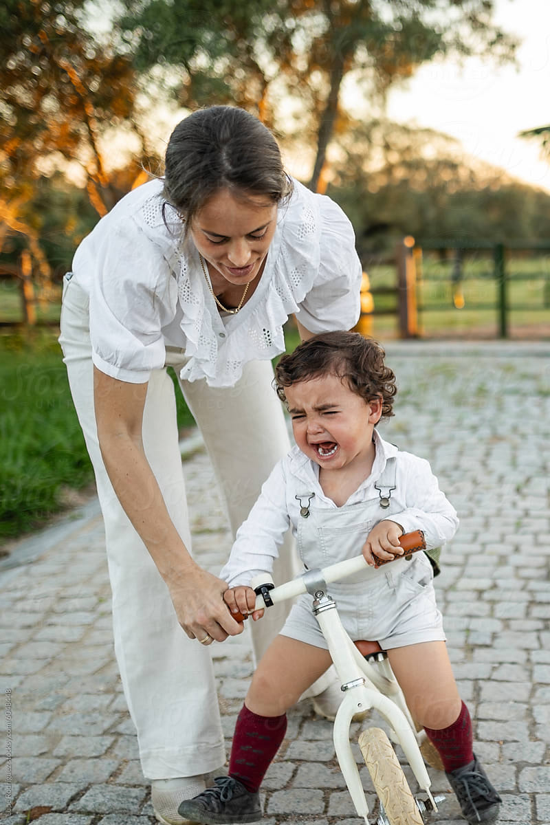 crying boy riding a bicycle in the garden at home with his mother