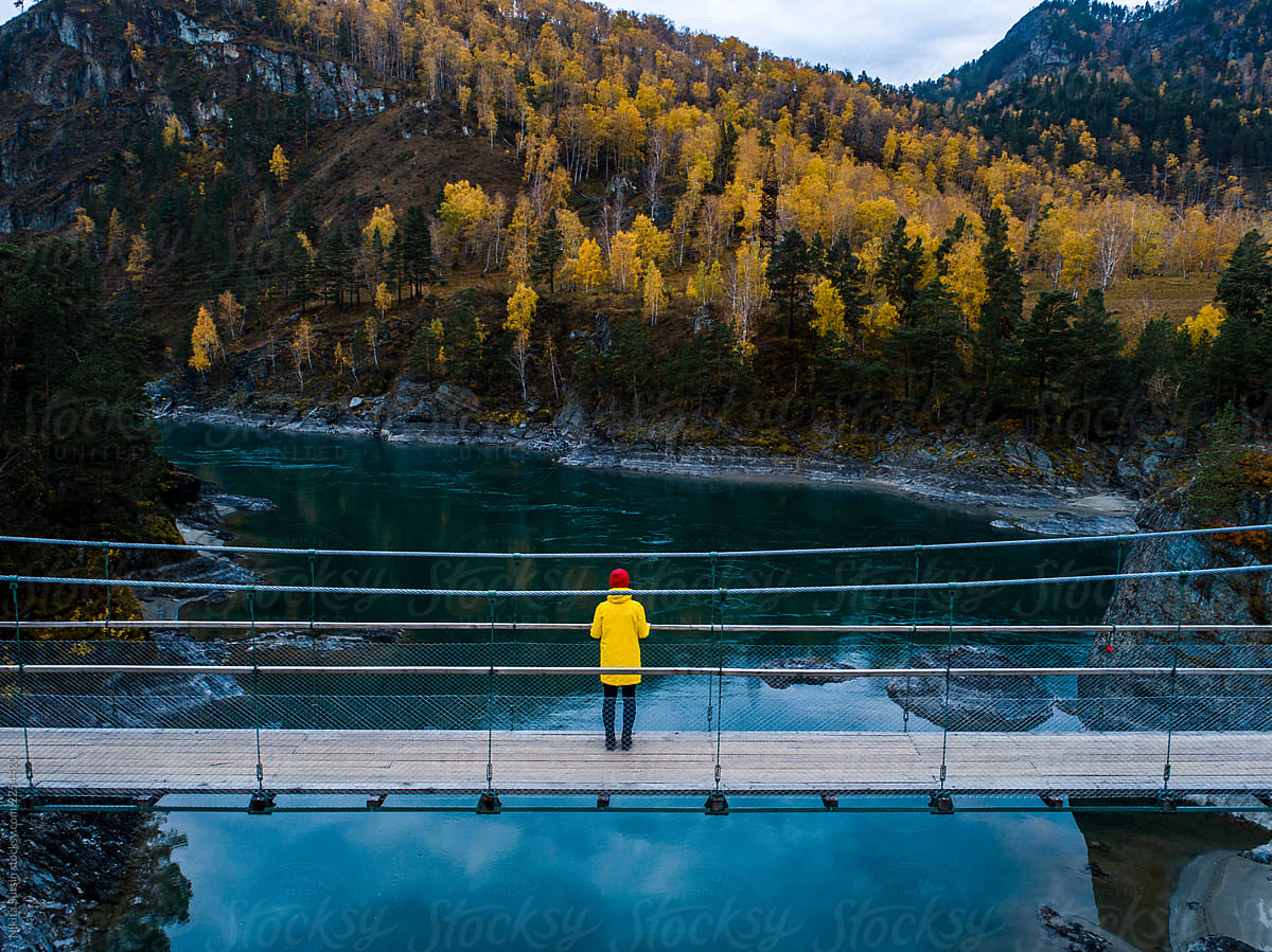 girl in a yellow jacket stands on a suspension bridge. Under the bridge flows a mountain river