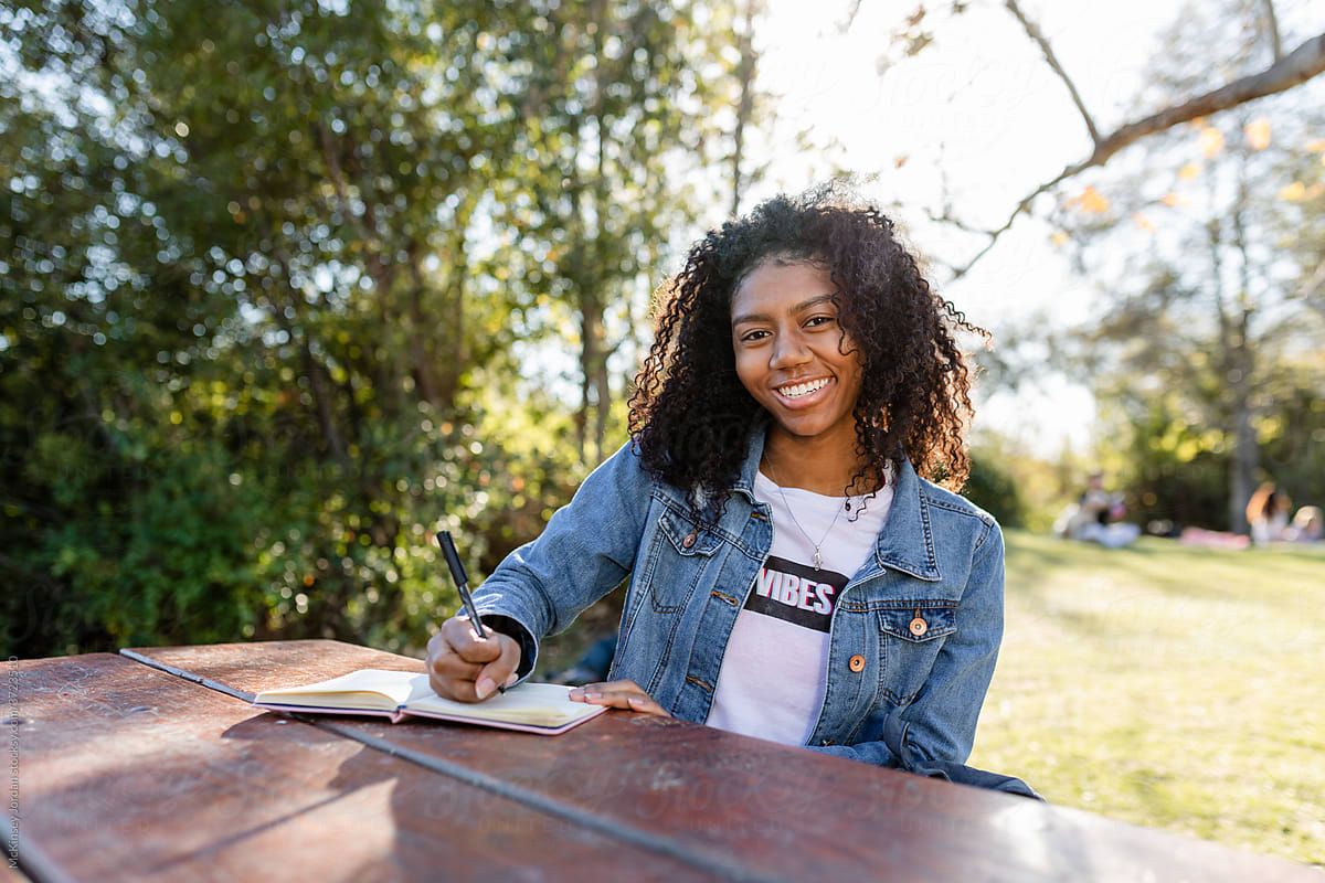 Young Woman Smiles at Camera While Writing at a Picnic Table in the Park