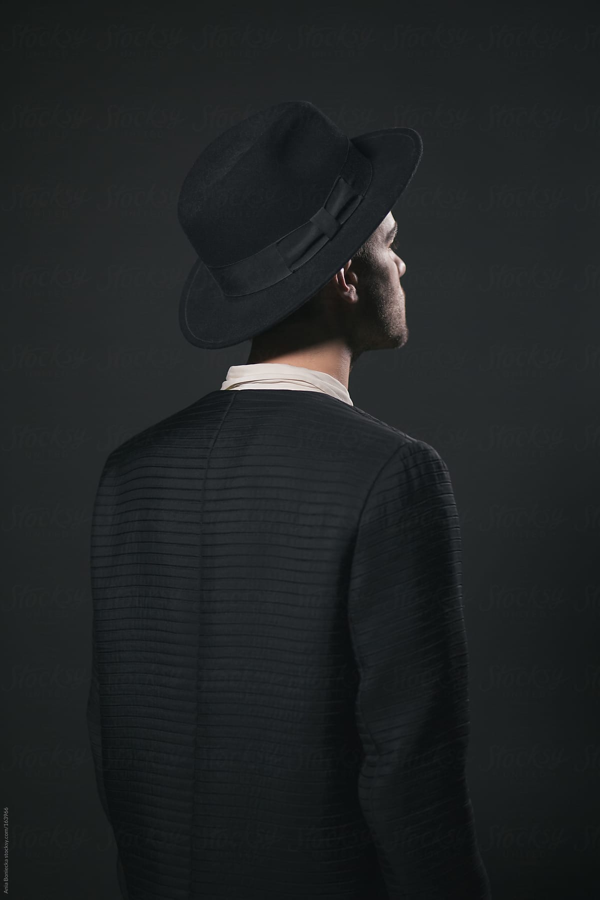 Dark portrait of the back of the man, wearing a hat against a black backgorund
