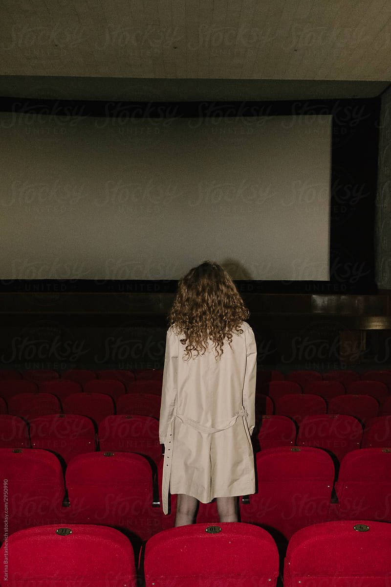curly-haired girl stands facing the cinema screen between rows of red chairs