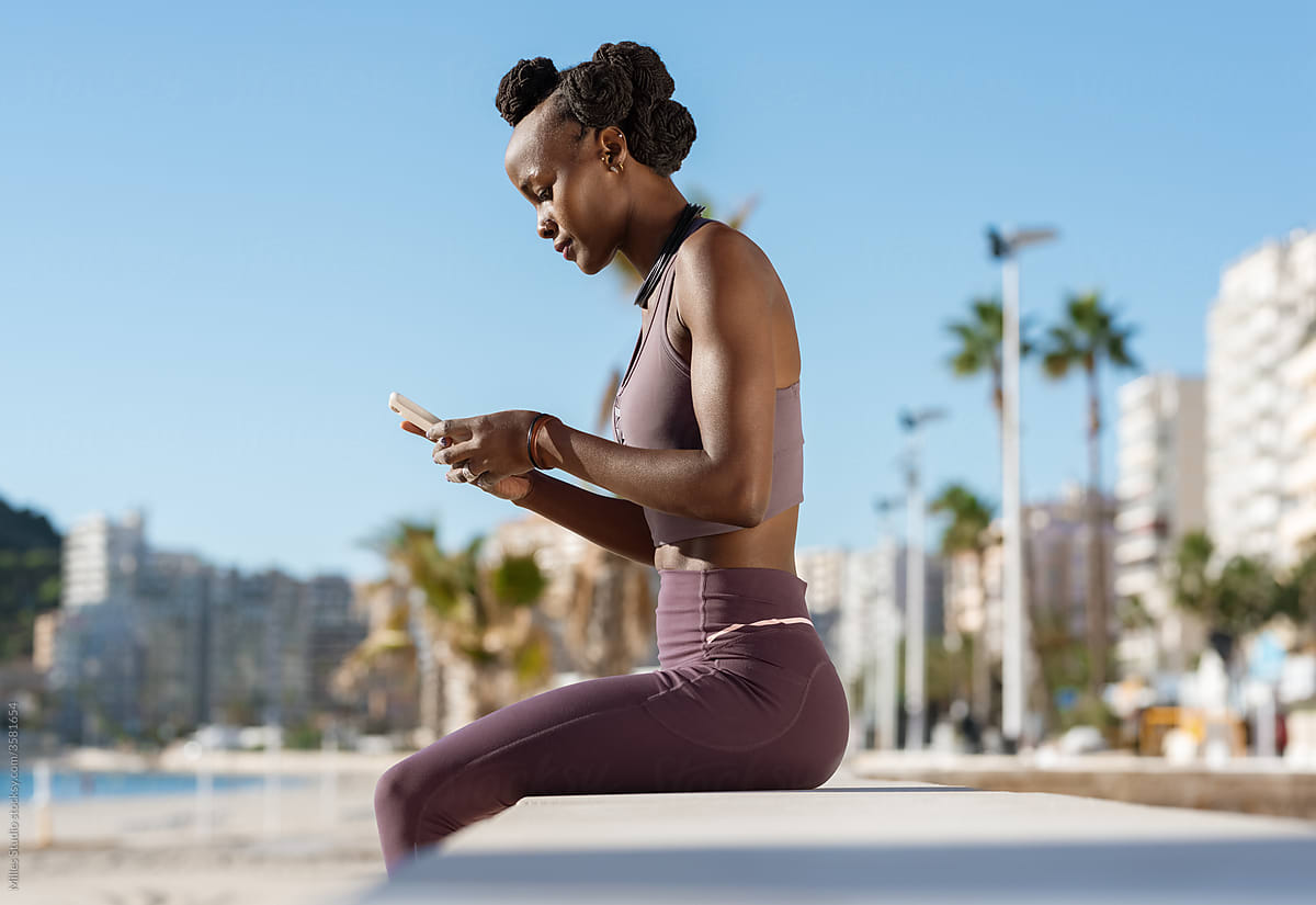 Black sportswoman sitting on border and using cellphone