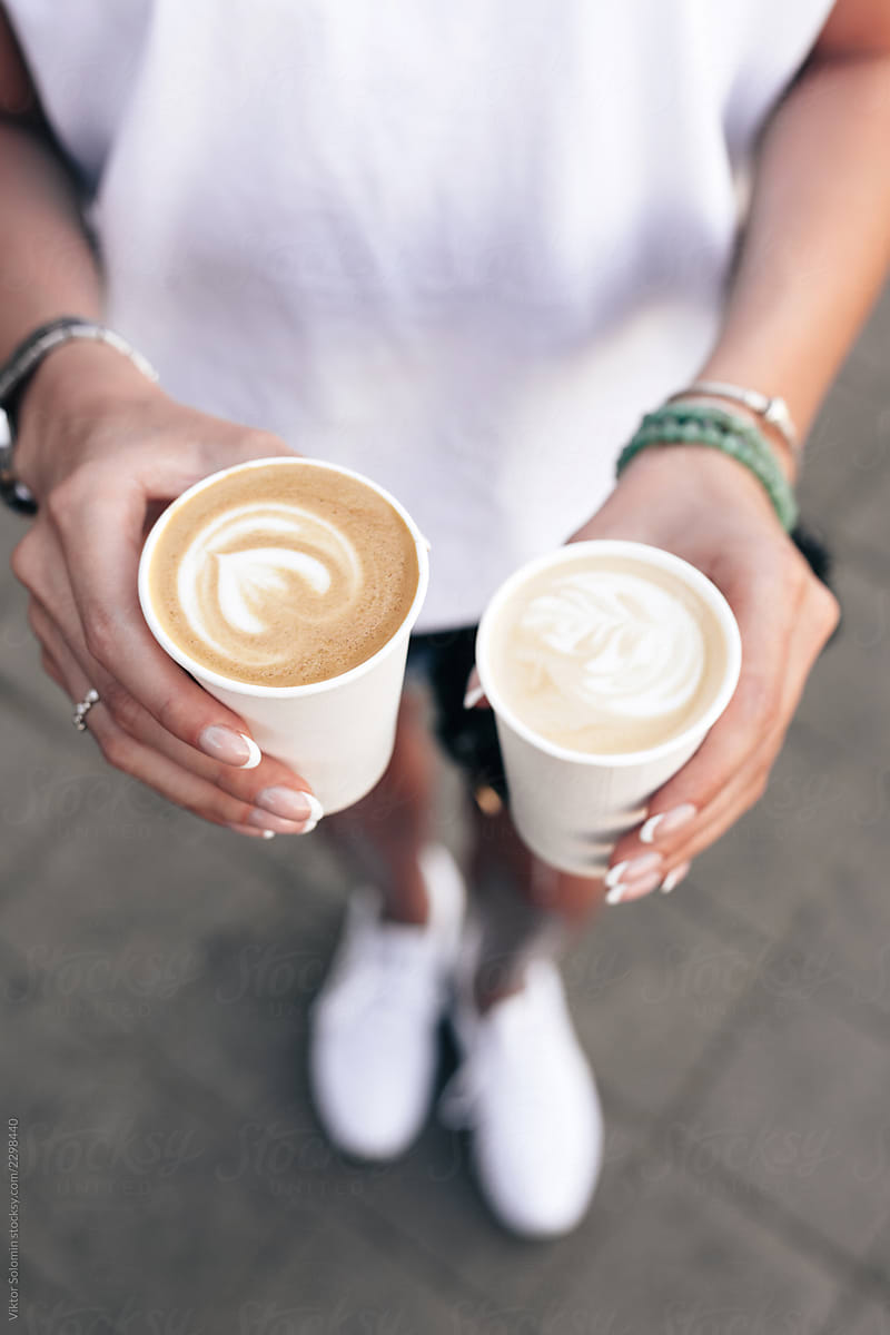 Summer street closeup of young beautiful hands holding two takeaway paper cups with cappuccino